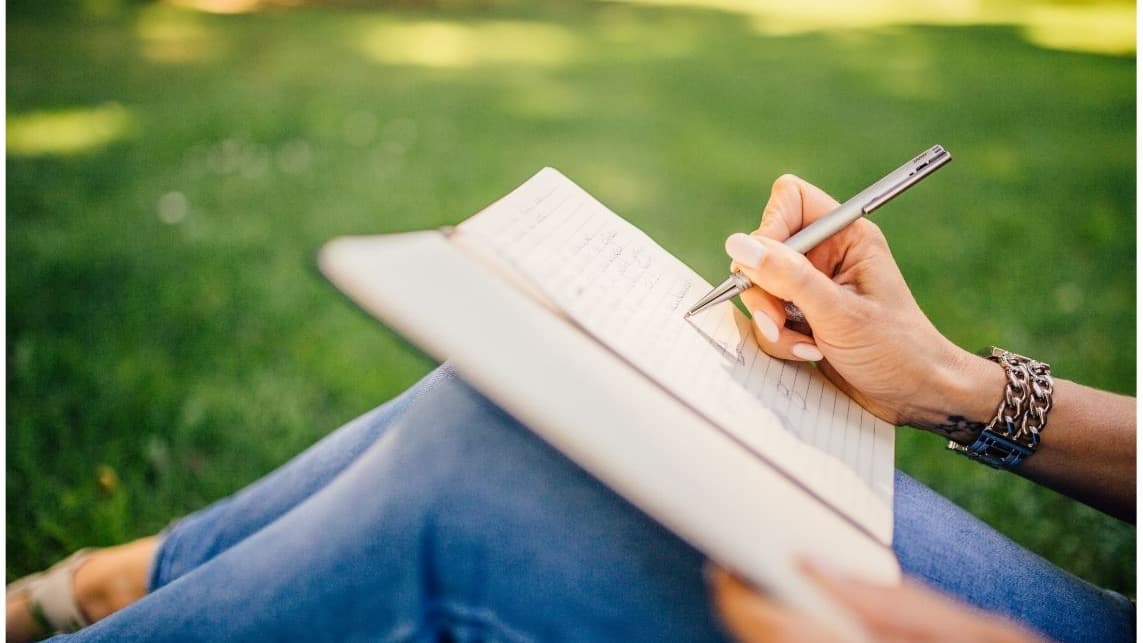 Hand schreibt in ein Notizbuch, im Hintergrund eine sonnige Wiese